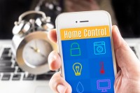 Hand holding smart phone with home control application with clock and computer at background, Smart home concept.